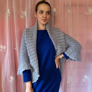 May include: A light gray crocheted cardigan with a shawl collar and bell sleeves. The cardigan is worn over a royal blue dress.