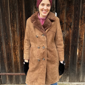 May include: A brown shearling coat with a double-breasted design. The coat features a sheepskin lining and a wide collar. The person is wearing a burgundy headband, black gloves, and blue jeans. The coat has silver buttons.