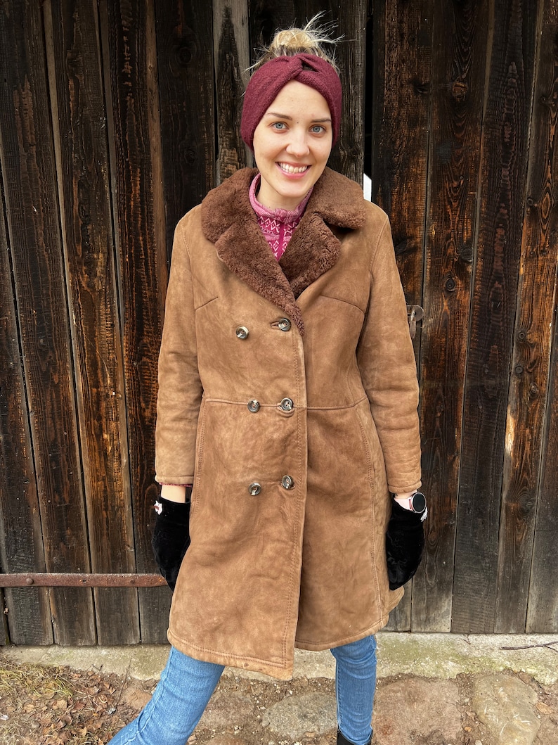 May include: A brown shearling coat with a double-breasted design. The coat features a sheepskin lining and a wide collar. The person is wearing a burgundy headband, black gloves, and blue jeans. The coat has silver buttons.