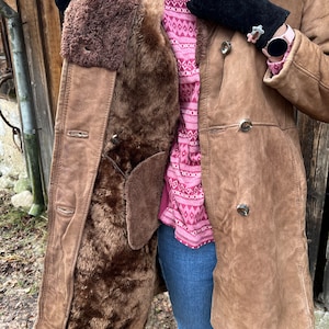 May include: A brown shearling coat with a button closure. The coat has a dark brown fur lining and a suede exterior. The person is wearing a pink patterned sweater, blue jeans, and black gloves.