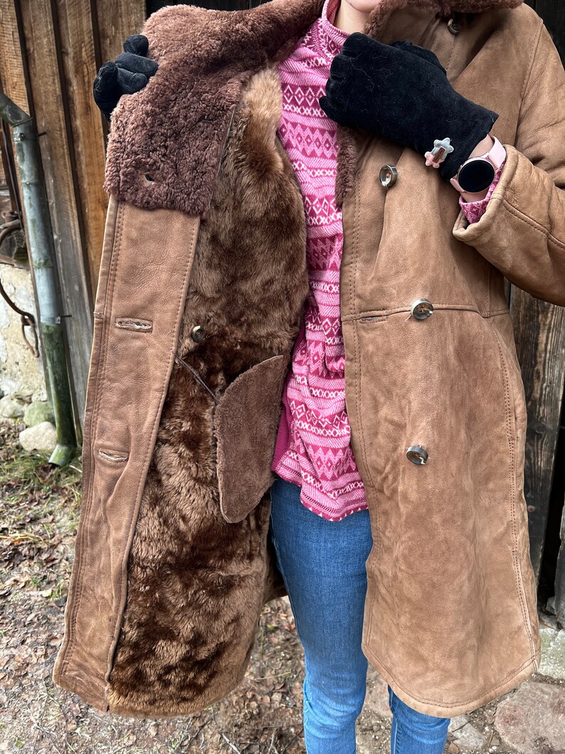 May include: A brown shearling coat with a button closure. The coat has a dark brown fur lining and a suede exterior. The person is wearing a pink patterned sweater, blue jeans, and black gloves.
