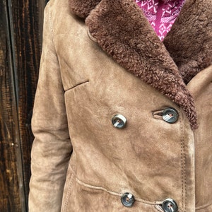 May include: A tan suede coat with a brown shearling collar and trim. The coat features a double-breasted design with large, decorative buttons. The coat is a classic style, suitable for cold weather.