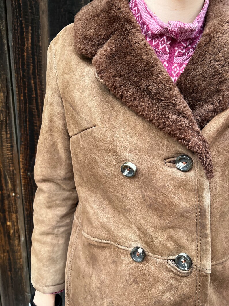 May include: A tan suede coat with a brown shearling collar and trim. The coat features a double-breasted design with large, decorative buttons. The coat is a classic style, suitable for cold weather.