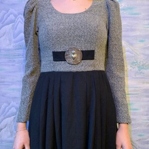 May include: A grey dress with a black skirt. The dress has long sleeves, a round neckline, and a black belt with a silver heart-shaped buckle. The skirt is pleated.