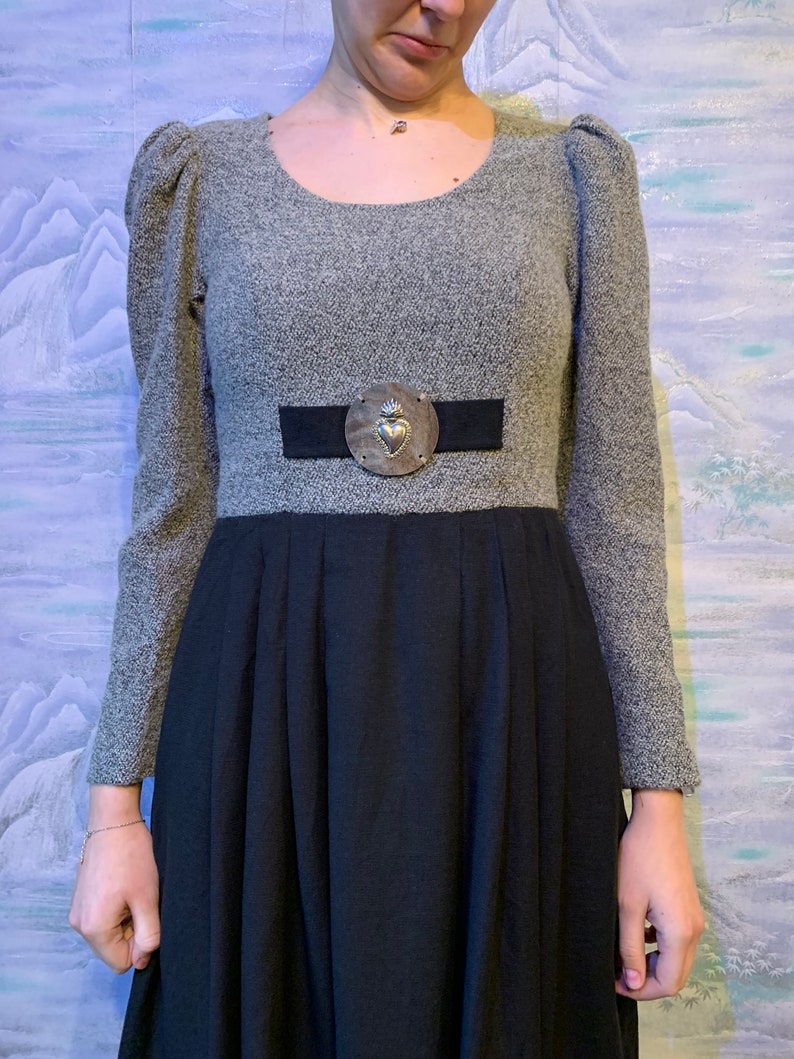 May include: A grey dress with a black skirt. The dress has long sleeves, a round neckline, and a black belt with a silver heart-shaped buckle. The skirt is pleated.