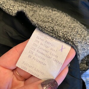 May include: A close-up of a fabric label showing the composition of the outer fabric and lining. The label lists percentages of wool, linen, cotton, viscose, and polyester. The lining is 100% viscose. The fabric is grey and black.