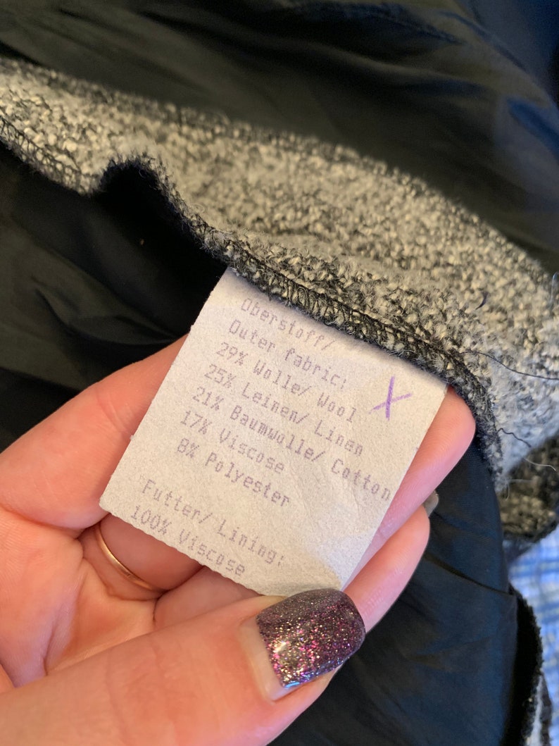 May include: A close-up of a fabric label showing the composition of the outer fabric and lining. The label lists percentages of wool, linen, cotton, viscose, and polyester. The lining is 100% viscose. The fabric is grey and black.