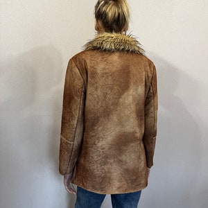 May include: A brown jacket with a textured, mottled finish and a tan faux fur collar. The jacket features long sleeves and a straight hemline, worn over blue denim jeans. The garment displays a weathered appearance with visible dark brown lines.