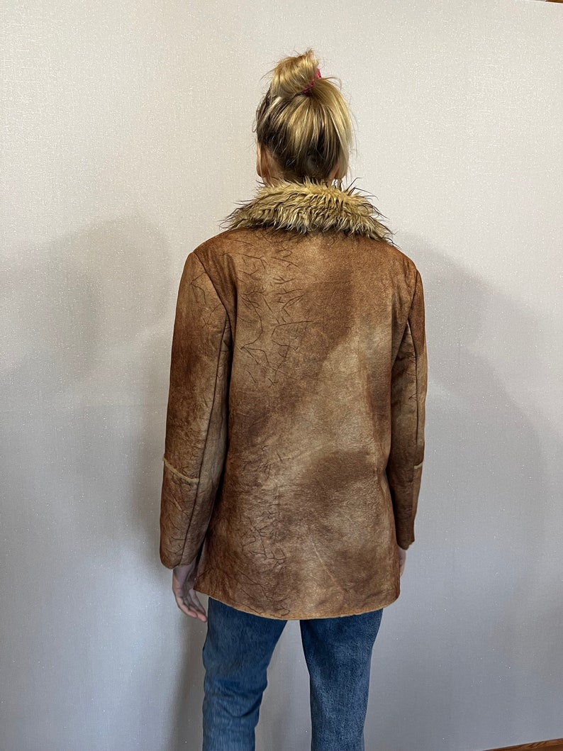 May include: A brown jacket with a textured, mottled finish and a tan faux fur collar. The jacket features long sleeves and a straight hemline, worn over blue denim jeans. The garment displays a weathered appearance with visible dark brown lines.