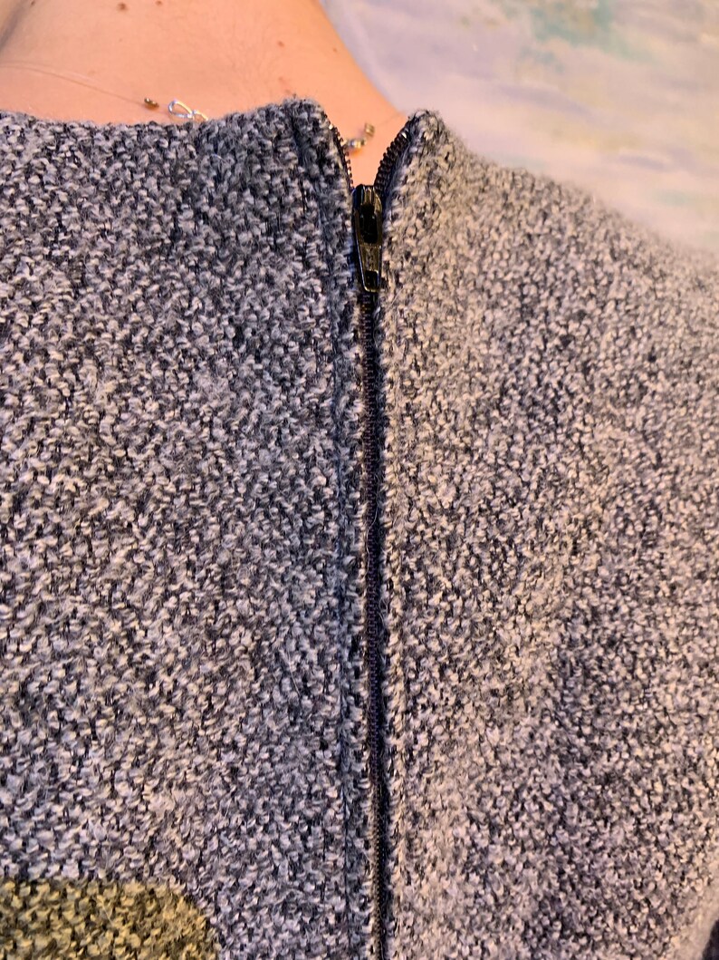 May include: Close-up of a textured grey and white fabric with a black zip down the centre. The fabric appears to be a woven material, possibly part of a jacket or coat.