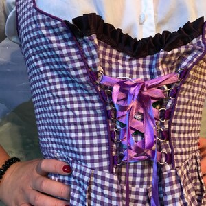 May include: A purple and white checkered bodice with a purple ribbon corset. The bodice has a black ruffled trim and is worn over a white blouse. The garment is a traditional style, possibly for a costume or themed event.