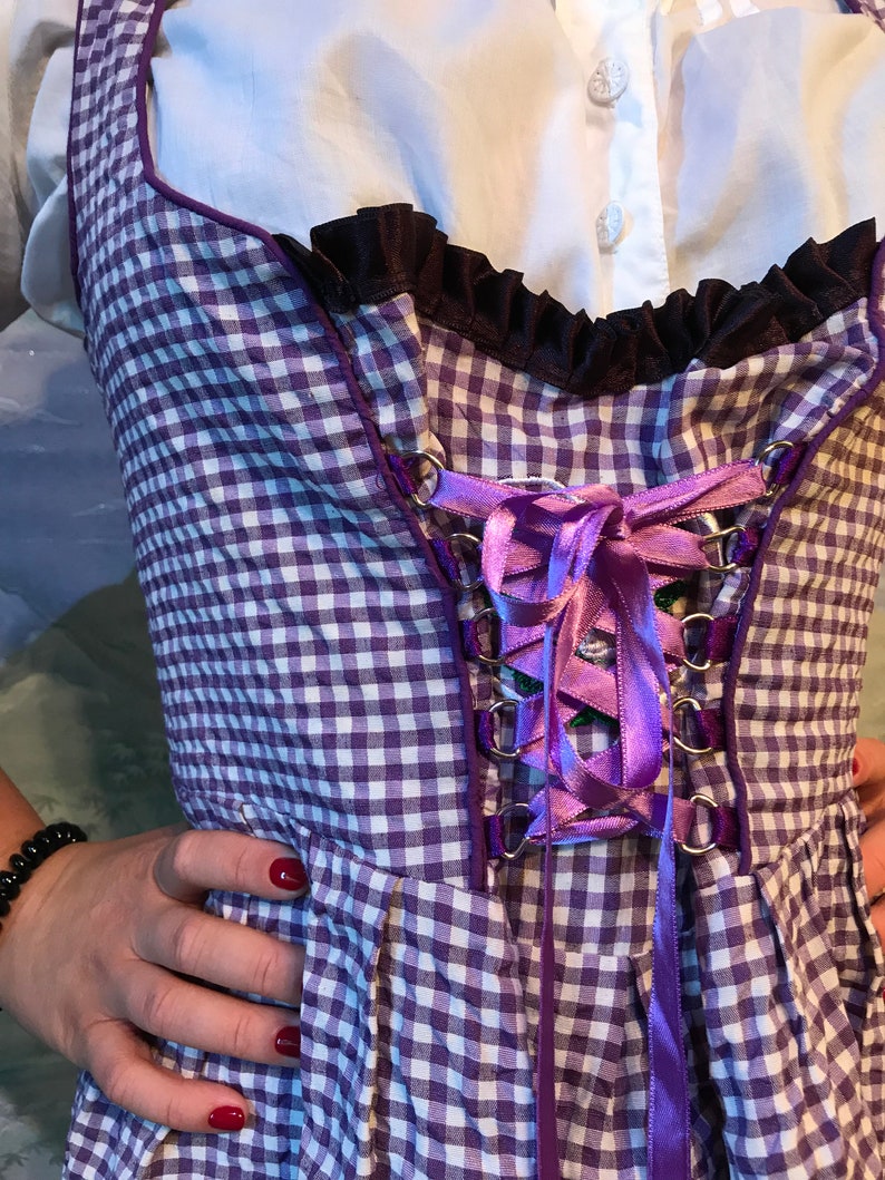 May include: A purple and white checkered bodice with a purple ribbon corset. The bodice has a black ruffled trim and is worn over a white blouse. The garment is a traditional style, possibly for a costume or themed event.