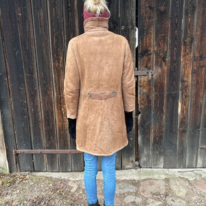 May include: A tan suede coat with a high collar and two decorative buttons at the waist. The coat is worn with blue jeans and black boots. The person is standing in front of a weathered wooden door.