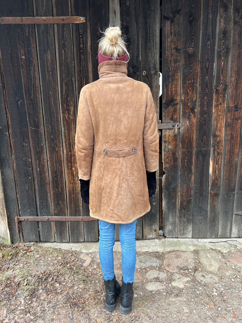 May include: A tan suede coat with a high collar and two decorative buttons at the waist. The coat is worn with blue jeans and black boots. The person is standing in front of a weathered wooden door.