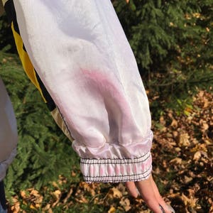 May include: Close-up of a white and yellow jacket sleeve. The sleeve has a pink stain and a black and pink striped cuff. The jacket is made of a shiny material.