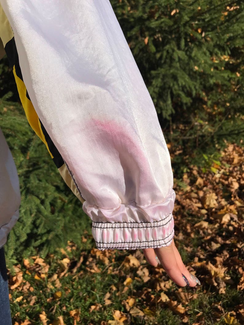 May include: Close-up of a white and yellow jacket sleeve. The sleeve has a pink stain and a black and pink striped cuff. The jacket is made of a shiny material.