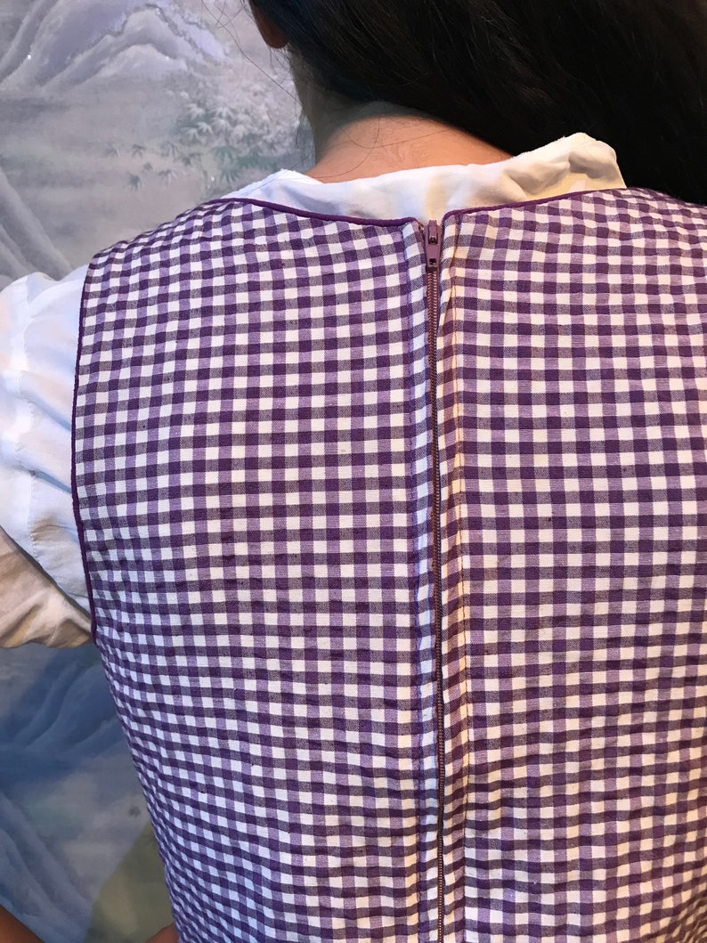 May include: A purple and white checkered vest with a full-length zipper down the back. The vest is worn over a white collared shirt. The zipper is a matching purple color.