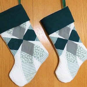 May include: Two Christmas stockings with a patchwork design. The top of each stocking is a solid dark green, with a green ribbon loop for hanging. The patchwork includes gray, white, and green patterned fabrics. The toe of each stocking is white.