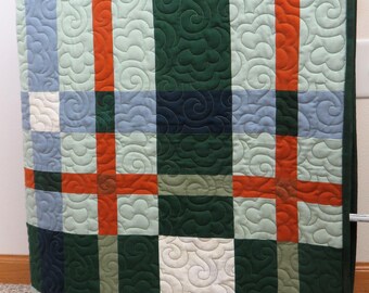 May include: A quilted throw blanket with a plaid pattern in shades of green, blue, orange, and white. The blanket is quilted with a swirling pattern.