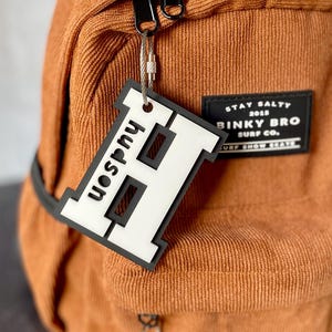 May include: A white and black personalized letter H keychain with the name Hudson on a brown corduroy backpack with a black patch that says "Stay Salty 2015 Binky Bro Surf Co."