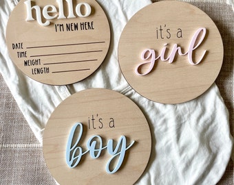 May include: Three wooden baby announcement signs. One sign says "hello I'm new here" with space for a date, time, weight, and length. Another sign says "it's a girl" with the word "girl" in pink. The last sign says "it's a boy" with the word "boy" in blue.