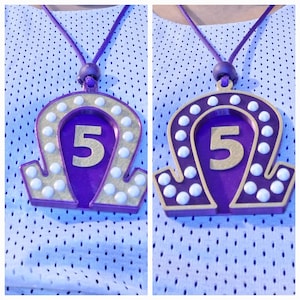 May include: Purple and gold necklace with a white number 5 in the center of a horseshoe-shaped pendant. The pendant is surrounded by white beads.