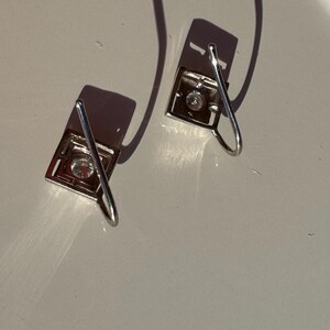 May include: A pair of silver earrings with a square design and a clear crystal in the center. The earrings have a simple, elegant design.