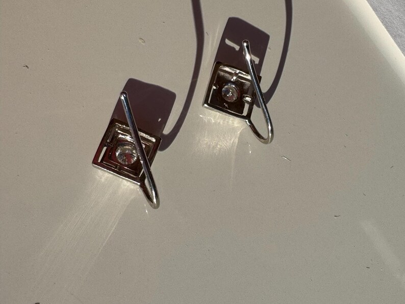 May include: A pair of silver earrings with a square design and a clear crystal in the center. The earrings have a simple, elegant design.