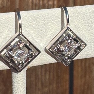 May include: A pair of silver dangle earrings with a square-shaped design. Each earring features a small, clear gemstone in the center.