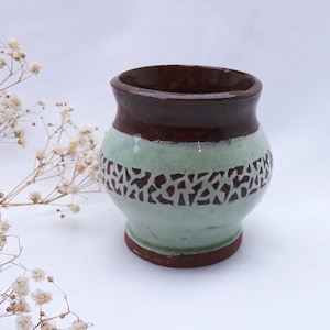 Peut inclure: Un petit pot en céramique brun et vert avec un motif décoratif autour du milieu. Le pot est verni et a une forme arrondie.