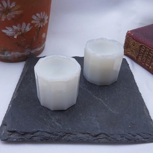 May include: Two white, octagonal candle holders on a black slate surface.