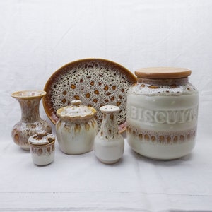 Puede incluir: Un juego de vajilla de cocina de cerámica marrón y blanca con un esmalte moteado. El juego incluye un jarrón, un tarro pequeño, un salero, un azucarero, un tarro grande con la etiqueta "BISCUITS" y una fuente.