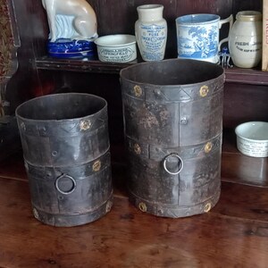 Könnte beinhalten: Zwei schwarze Metallbehälter mit Messingakzenten und einem Ringgriff. Die Behälter stehen auf einer Holzoberfläche.