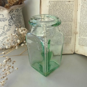 Peut inclure: Un bocal en verre carré vintage avec une teinte vert clair. Le bocal a une large ouverture et une surface légèrement texturée. Des fleurs séchées et un livre ouvert sont en arrière-plan, créant une esthétique rustique.