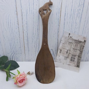 May include: A wooden spoon with a carved bear on the handle. The spoon is brown and has a worn look. It is sitting on a white surface.