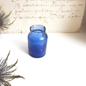 Puede incluir: Un pequeño tarro de vidrio azul cobalto con una superficie ligeramente texturizada. El tarro tiene un cuerpo redondeado y un borde ensanchado e irregular. El fondo incluye cardo seco y un trozo de papel envejecido con escritura descolorida.