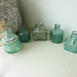 Peut inclure: Cinq flacons d'encre en verre vintage de formes et de tailles différentes. Les flacons sont transparents et ont une teinte vert clair. Ils sont disposés en rangée sur une surface blanche.