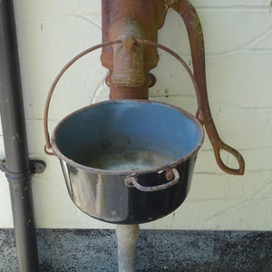 Puede incluir: Una bomba de mano oxidada con una cuenca de metal negro unida a ella. La bomba está montada en una pared blanca.
