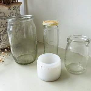 Może przedstawiać: Kolekcja zabytkowych szklanych słoików i pojemników. Obejmuje wysoki, przezroczysty słoik, mały słoik z żółtą pokrywką, krótki, szeroki słoik i mały biały pojemnik. Słoiki są ułożone na białej powierzchni.