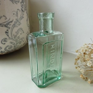 Può includere: Una piccola bottiglia di vetro verde chiaro di forma quadrata con la parte superiore arrotondata. La bottiglia ha la parola "HINDS" in rilievo sul lato.