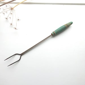 Könnte beinhalten: Eine antike Metall-Bratgabel mit einem türkisfarbenen Griff. Der Griff ist zylindrisch und weist Gebrauchsspuren auf, mit einem Metallband am Fuß. Die Gabel hat zwei Zinken und einen langen, schlanken Metallschaft. Der Hintergrund ist weiß.