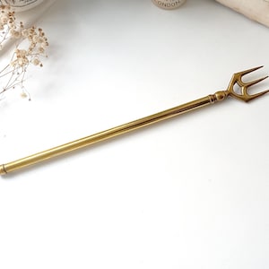 Könnte beinhalten: Ein goldfarbener Brieföffner in Form eines Dreizacks. Der Brieföffner hat einen langen, schlanken Griff und eine dreizackige Gabel an der Spitze. Der Dreizack liegt auf einer weißen Oberfläche.