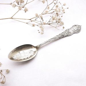 May include: A silver-toned metal souvenir spoon featuring an ornate, embossed handle with floral and shell motifs. The bowl of the spoon displays an engraved illustration of a building and the text "COURT-HOUSE" in clear, dark lettering.