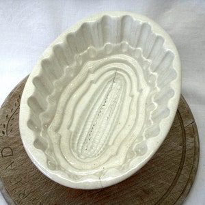 Puede incluir: Un molde de cerámica blanca para hornear un pan de maíz. El molde tiene un borde acanalado y un diseño detallado de mazorca de maíz en el centro.