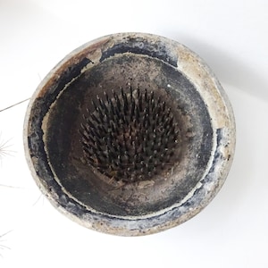 Op de afbeelding: Een vintage keramische bloemenkruk met een zwarte en bruine glazuur. De kruk heeft een ronde basis met een midden dat is gevuld met metalen pinnen om bloemstengels vast te houden.