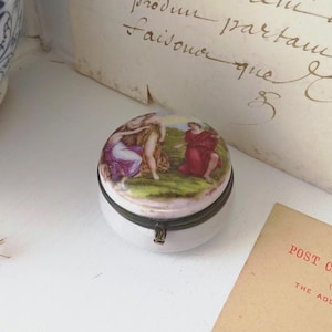 Peut inclure: Une petite boîte à bijoux ronde en porcelaine avec une fermeture métallique à charnière. Le couvercle présente une scène peinte colorée représentant trois personnages dans un paysage. La base est blanche et le bord métallique est vieilli.