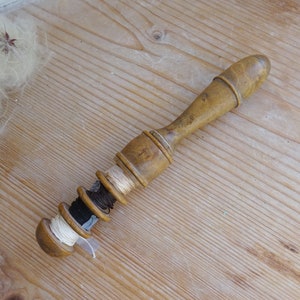 Op de afbeelding: Een vintage houten draadhouder met meerdere spoelen draad in verschillende kleuren, waaronder wit, beige, zwart en bruin. De houder heeft een afgeronde handgreep en een spoelhoudergedeelte. Het item is geplaatst op een lichtgekleurd houten oppervlak.