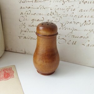 May include: A vintage wooden salt shaker with a rounded body and a cap with several small holes. The shaker is a warm brown color and sits on a white surface. A vintage postcard and handwritten document are in the background.