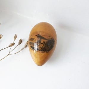 May include: A vintage, egg-shaped wooden container with a landscape image. The container is light brown with a darker image of a building and text. The egg is cracked at the top. The background is white.