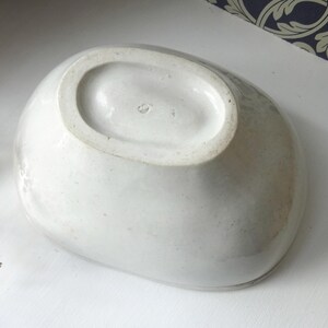 May include: A white, oval-shaped ceramic bowl with a slightly textured surface. The bowl has a wide rim and a smooth interior. The underside of the bowl is visible, showing the base and a small marking. The bowl is likely vintage.